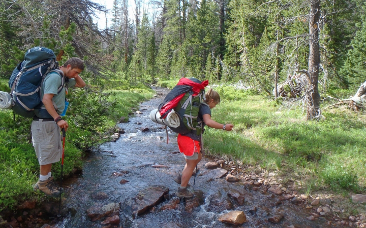 A person wearing a backpack crosses a creek in ankle-deep water while another person watches